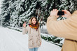 © Elizaveta - Young couple in love rest in the park winter. A man holds mobile phone in hands and takes a photo of girlfriend against the background of snowcovered trees. Woman in jacket and red backpack