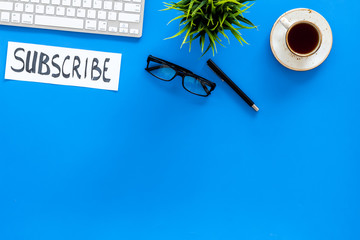 Poster - Email subscribe concept. Hand lettering subcribe on work desk with plant, glasses, computer keyboard, cup of coffee on blue background top view copy space