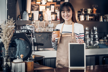 Poster - Asian female barista wear jean apron holding coffee cup and blank blackboard coffee menu at counter bar with smile face,cafe service concept,business owner start upLeave space for adding your text.