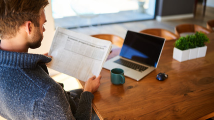 over the shoulder image of an attractive young man holding a newspaper while seated at his beautiful wooden dining table at home, with a hot cup of tea and his laptop ready to work.