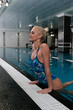 Attractive young blonde woman climbs out of the water in the pool, leaning on the side and looking forward