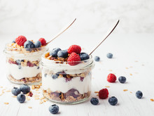 Two Jars With Tasty Parfaits Made Of Granola, Berries And Yogurt On White Wooden Table. Shot At Angle.