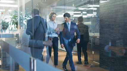 businessman and businesswoman walking through glass hallway, discussing work and using tablet comput