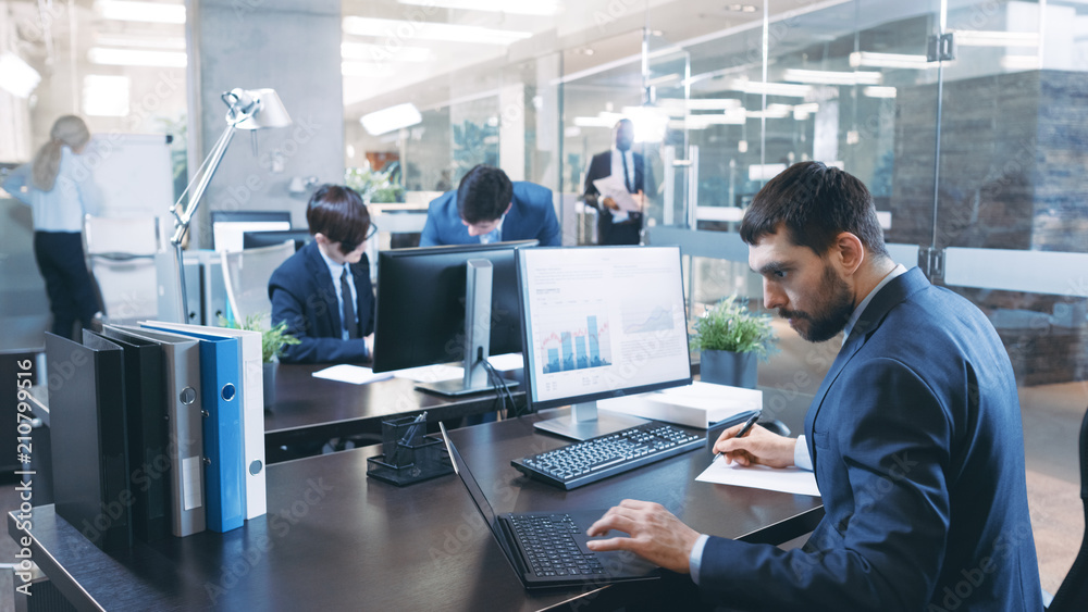 Professional Businessman Works on His Desktop Computer, Uses Laptop. In the Background Busy Office with Diverse Group of Business People.  Modern Glass and Marble Corporate Building.