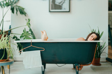 Tilt Shift Portrait Of Young Beautiful Girl In Pink Dress Lying In Empty Vintage Cast-iron Bath Inside Decorative In French Style Room With Green Plants. Odd Unusual Strange Woman Home Relaxation.