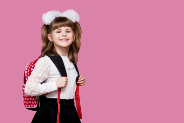 cute smiling little girl in school uniform and white bows with backpack on pink background. back to 