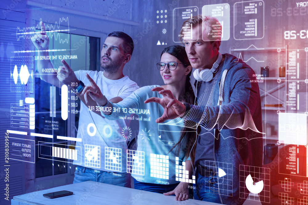 Busy workers. Clever professional web developers standing together and carefully touching a holographic screen while working in their modern office