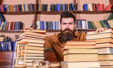 Leinwandbilder - Teacher or student with beard sit at table with hourglass and glasses, defocused. Man on strict face sit between piles of books, while studying in library, bookshelves on background. Bookworm concept.