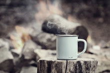 Cup Of Hot Steaming Coffee Sitting On An Old Log By An Outdoor Campfire With A Vintage Antique Edit. Extreme Shallow Depth Of Field With Selective Focus On Mug.
