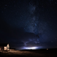 a wood cabin with the porch light on with a wooden fence and grass field with a lightning storm and 