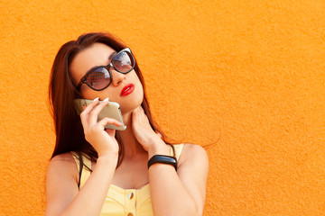 Wall Mural - Fashion portrait of pretty smiling woman in sunglasses talking on the smartphone against the orange wall.fitness bracelet on the hand