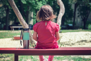 a little upset girl in a red dress sits on a bench alone