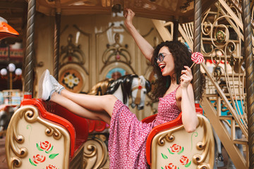 Cheerful lady with dark curly hair in sunglasses and dress holding lolly pop candy in hand and riding on carousel while spending time in amusement park