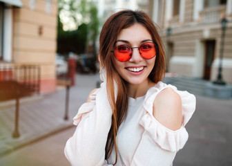 Wall Mural - Portrait Happy young adult woman smiling with teeth smile and long brunette hair outdoors ,walking on city street at sunset time wearing trendy sunglasses,beige shirt and stylish bag.Toned,copy space.