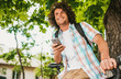 © iuricazac - Bottom view of happy young male student smiling with curly hair wearing shirt with backpack, sitting on the bike on the street, relaxing, texting messages on smart phone. People and education