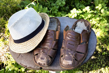 A Fedora Hat Over Old Brown Sandals On A Grey Garden Table