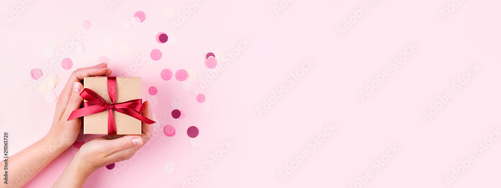 Woman's hands holding kraft gift box with red bow on pink background decorated with confetti.. Top view, holiday present concept with copy space, banner. - obrazy, fototapety, plakaty 
