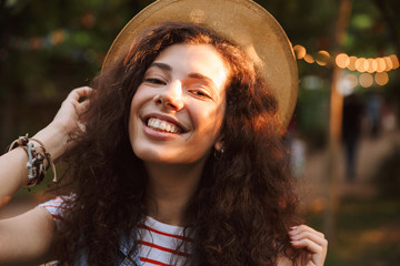 Poster - Image closeup of beautiful young woman 18-20 wearing summer straw hat smiling with closed eyes, while walking through park in sunny day with trees background