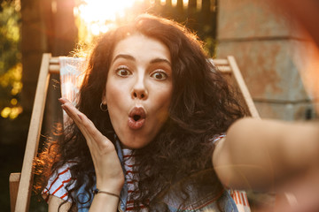 Sticker - Picture of attractive joyful girl 18-20 looking at camera emotionally, while taking selfie photo during rest in lounge chair in park on sunny day