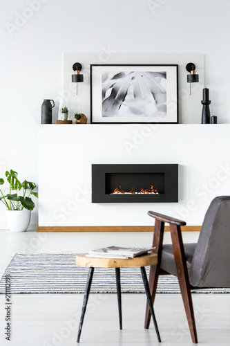 table next to grey armchair in white loft interior with poster abovetable next to grey armchair in white loft interior with poster above black fireplace real photo