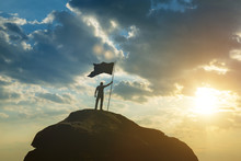 Silhouette Of A Man On Top Of A Mountain With The Flag Of Victory. Against The Background Of The Sky In The Sunset. High Achievements, Success In Business, Leadership,