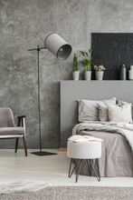 Stylish Standing Lamp Next To A Bed In A Monochromatic Grey Bedroom Interior With A Footstool, An Armchair And Decorative Plants In Pots Against The Wall. Real Photo.