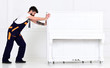 Man with beard and mustache, worker in overalls pushes piano, white background. Courier delivers furniture in case of move out, relocation. Courier service concept. Loader moves piano instrument.