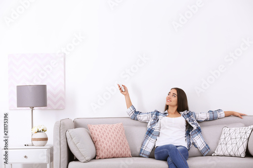 Fotografía Young woman switching on air conditioner while sitting on sofa near white wall