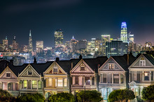 Backed By The Night Skyline Of The City Of San Francisco, California, The Victorian Era Houses Near Alamo Square Park, Are Painted In Colors To Accentuate Their Architectural Details.