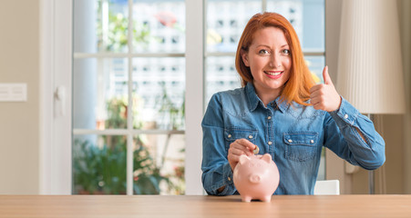 Sticker - Redhead woman saves money in piggy bank at home happy with big smile doing ok sign, thumb up with fingers, excellent sign