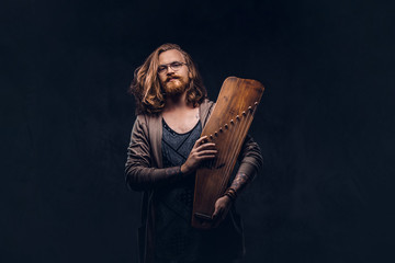 Wall Mural - Redhead hipster male with long luxuriant hair and full beard dressed in casual clothes holds a Russian traditional musical instrument - gusli. Isolated on the dark background.