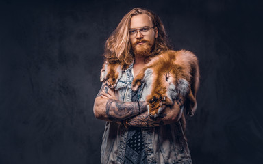 Wall Mural - Tattoed redhead hipster male with long luxuriant hair and full beard dressed in a t-shirt and jacket holds the fox skin on a shoulder in a studio. Isolated on the dark background.