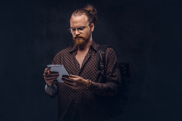 Wall Mural - Portrait of a redhead hipster student in glasses dressed in a brown shirt, holds a backpack and digital tablet, posing at a studio. Isolated on a dark background.