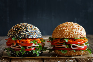 Two fresh appetizing hamburgers (dark and light) with sesame seeds, close-up, with fresh vegetables (tomato, sweet pepper), Korean carrots, parsley and ham. Black blurred background.