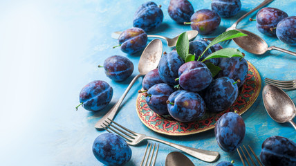 Wall Mural - Plums and leaves fresh arrangement on plate with forks and spoons on blue painted background and bright light in studio