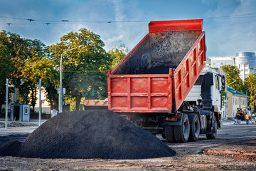 Transportation of asphalt to the site of road works. Heavy orange tipper unloads fresh hot  smoking asphalt mix on a prepared surface for paving. Building new roadway in the city.