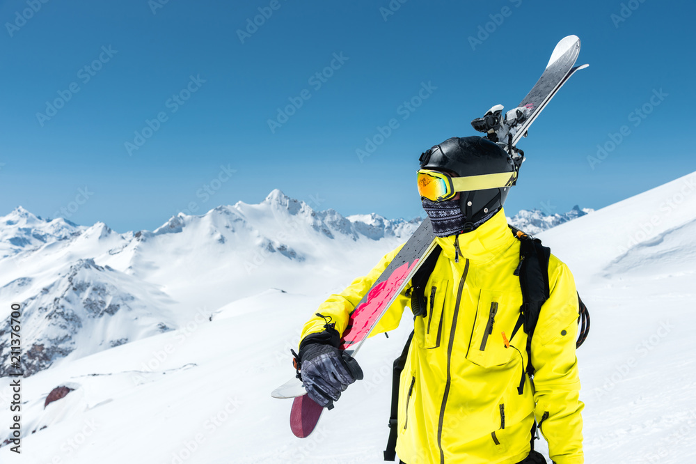 Obraz na płótnie A portrait of a skier in a protective helmet and glasses is a mask and scarf with skis on his shoulder in the snow-capped mountains of the Caucasus. Skiing, w salonie