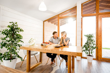 Young and happy couple having a breakfast sitting at the wooden table in the modern country house with big window on the background