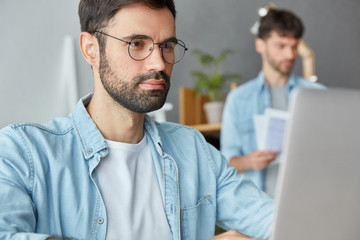 Wall Mural - Serious bearded young prospective male entrepreneur makes business research on laptop computer, looks attentively through information while his partner stands in background, studies company`s profit