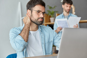 Wall Mural - Business and teamwork concept. Concentrated bearded young male startaper makes research or future project, looks through graphics on laptop computer, his colleague studies documentation in background