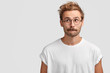 © wayhome.studio - Puzzled handsome male with mustache, bites lower lip and looks curiously aside, thinks about something, dressed in casual white t shirt, stands against blank wall with copy space for your text
