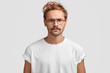 © wayhome.studio - Indoor shot of serious man with grumpy expression, being discontent with noisy neighbours, dressed in casual white t shirt and spectacles, poses indoor. Attractive hipster has strict look and mustache