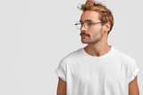 Fototapeta  - Profile shot of serious stylish hipster looks aside with confident expression, turns head aside, looks at something into distance, wears round spectacles, isolated over white background, blank space