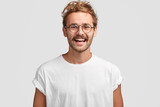 Fototapeta  - Horizontal shot of happy hipster male with toothy smile, wears casual white t shirt and glasses, being in good mood after unforgettable journey with girlfriend, isolated on white background.