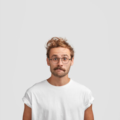 Wall Mural - Vertical shot of puzzled European male with curly haircut, purses lips and looks in bewilderment, surprised to see big sales, dressed in casual white t shirt, isolated over studio background