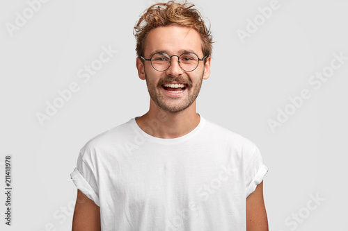 Cuadros en Lienzo Horizontal shot of happy hipster male with toothy smile, wears casual white t shirt and glasses, being in good mood after unforgettable journey with girlfriend, isolated on white background