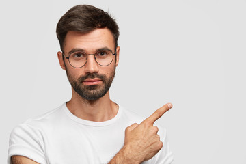 Wall Mural - Horizontal shot of attractive European male with serious expresssion, wears round glasses, shows blank space for your advertising content, suggests you to go in this place. People and advertisement
