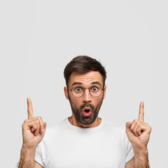 Wall Mural - Vertical shot of stunned bearded young male has frightened expression, indicates with both index fingers upwards, shocked to notice something above head, isolated on white wall. Monochrome concept