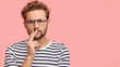 © wayhome.studio  - Horizontal shot of serious displeased man frowns face, keeps hand on mouth, has curly hair and stubble, wears striped t shirt, round spectacles, stands against pink background with copy space