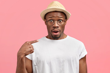 Wall Mural - Amazed handsome dark skinned male tourist in straw hat and white t shirt, points at himself with fore finger, has astonished expression, poses against pink studio background, lost in big city